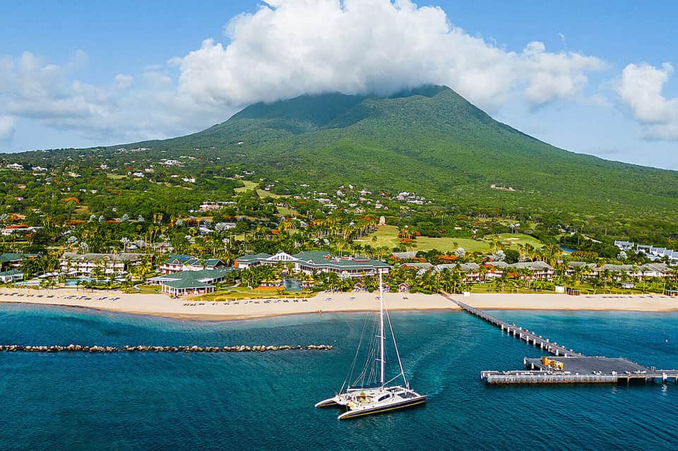 Sailboat anchored offshore with volcanic mountain, beach, and coastal town in background