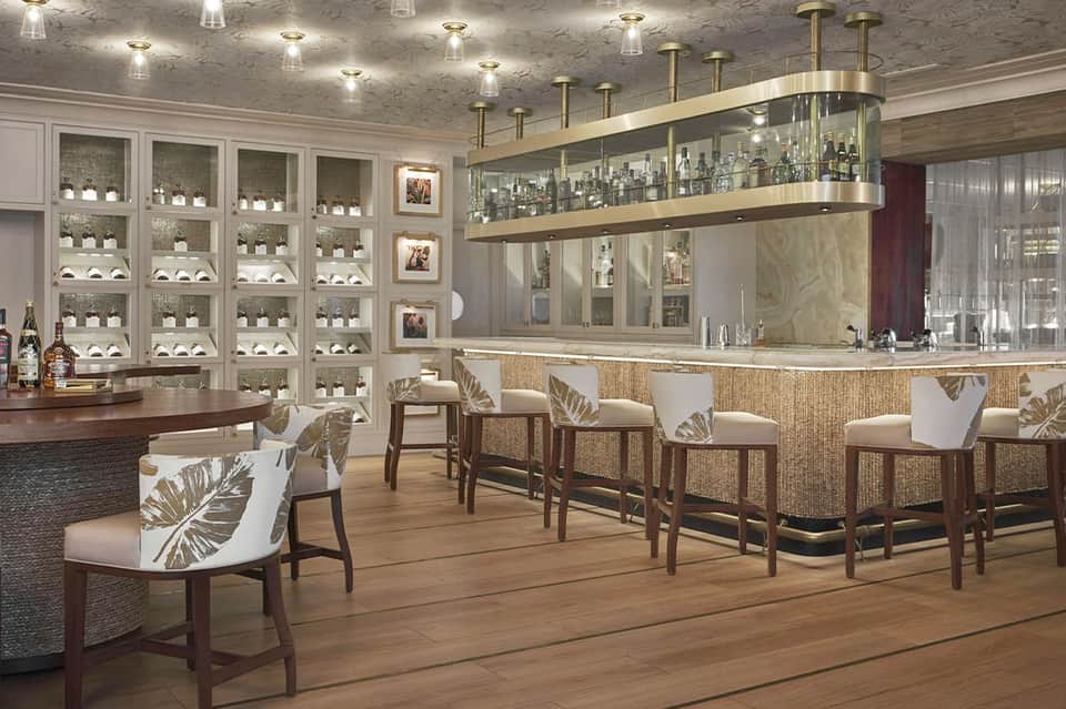 Luxury bar with illuminated shelving displaying bottles, woven bar seating, and tropical accents