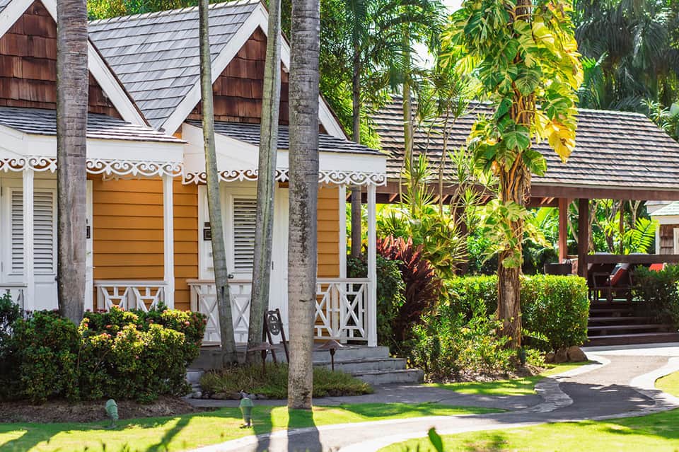 Tropical resort cottage with brown shingled roof, white trim, wooden doors, and lush green landscaping