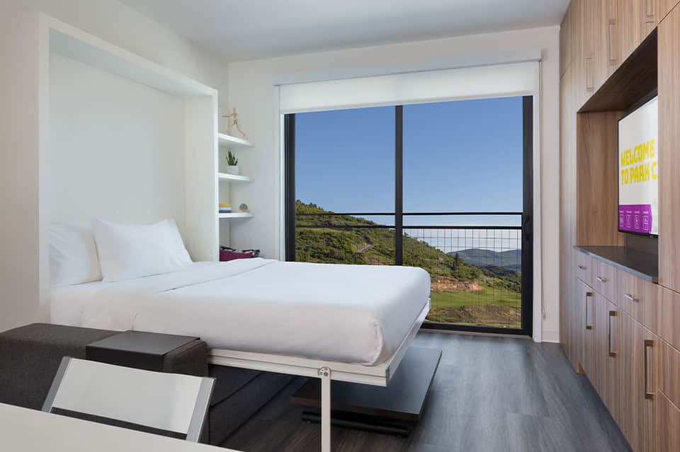 Modern hotel room with white bedding, balcony overlooking green hillside landscape