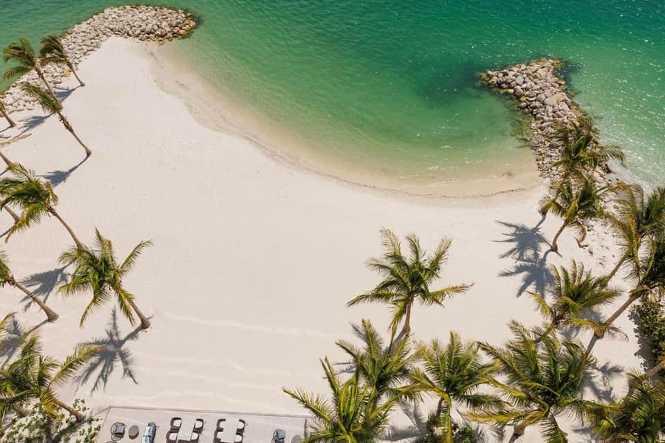 Aerial view of pristine beach with turquoise water, white sand, palm trees, and rocky jetties