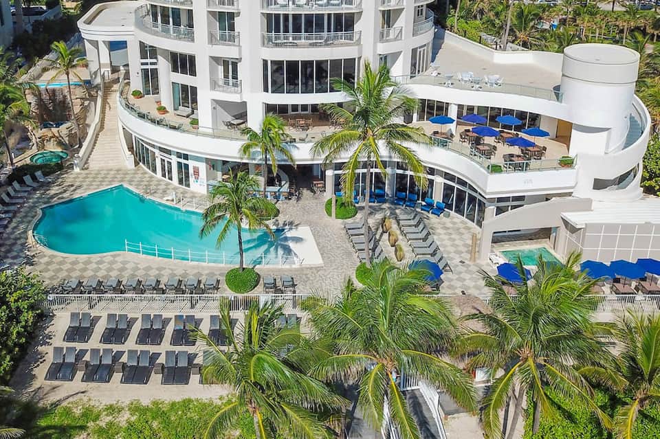 Aerial view of resort with curved white building, turquoise pool, palm trees, and blue umbrellas