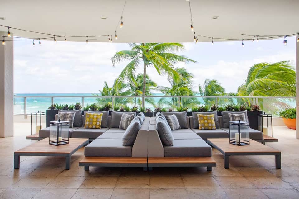 Modern terrace lounge with ocean view, grey seating, palm plants, and string lighting