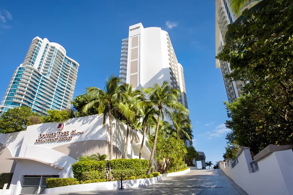Beachfront resort entrance with tall palm trees, white high-rise buildings, and manicured landscaping