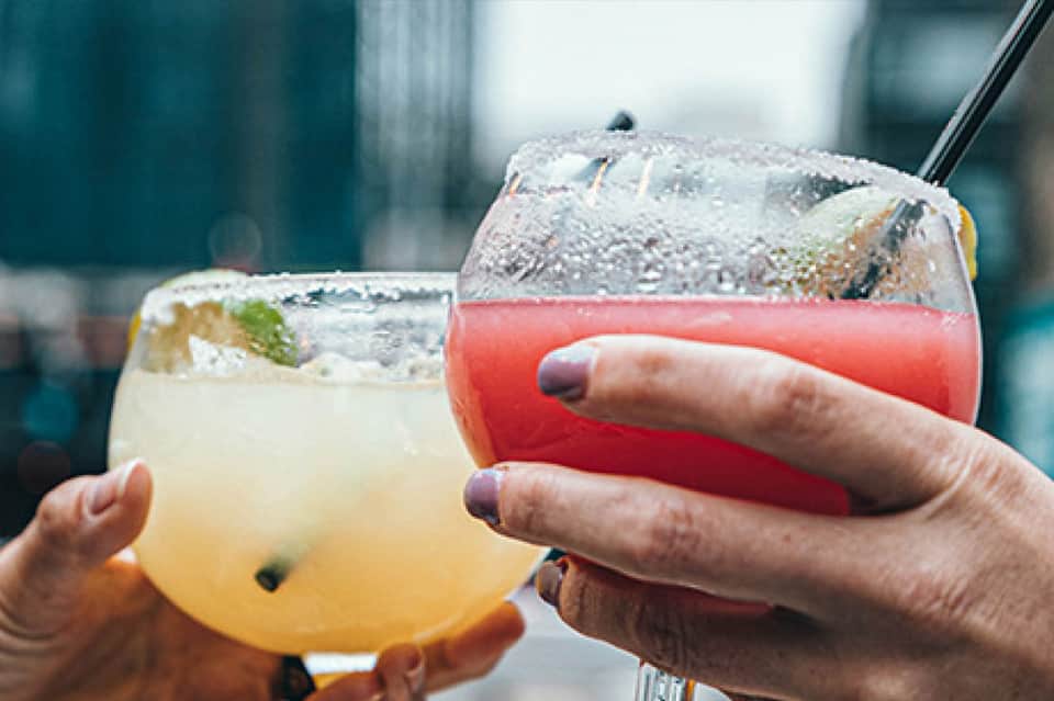 Two colorful tropical cocktails with ice and garnish held by person at bar