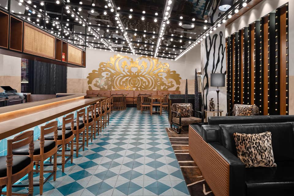 Stylish hotel bar with ornate gold wall art, teal checkered flooring, wooden chairs, and leopard print accents