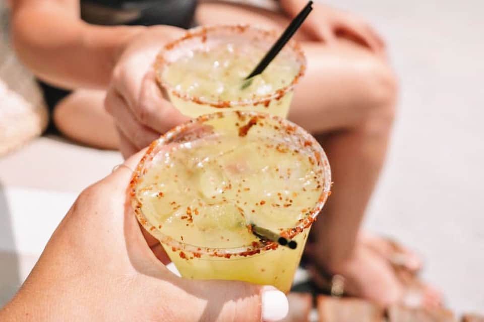Two spicy margarita cocktails with chili-salt rim and black straws held in hands by poolside