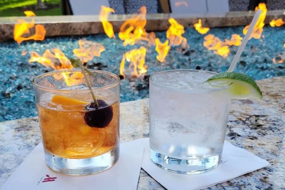 Two poolside cocktails on granite edge - amber drink and clear drink with fire pit and pool background