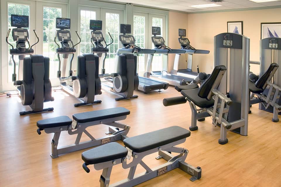 Bright fitness center with elliptical machines, treadmill, weight benches, and large windows overlooking gardens