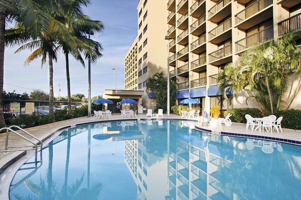 Resort pool area with blue umbrellas, palm trees, white loungers, and multi-story hotel building