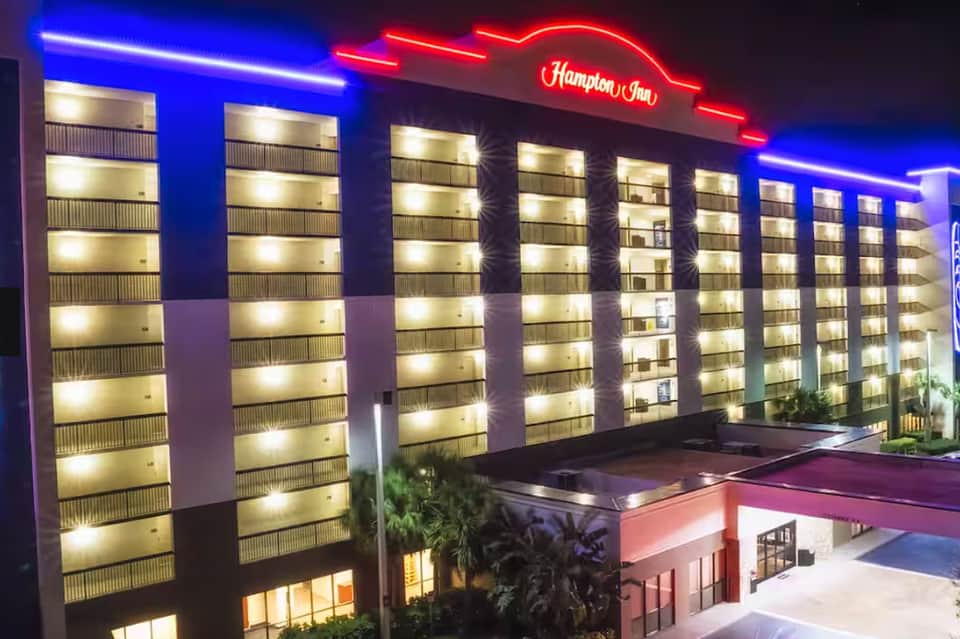 Hampton Inn hotel exterior at night with blue neon trim, illuminated signage, and palm trees