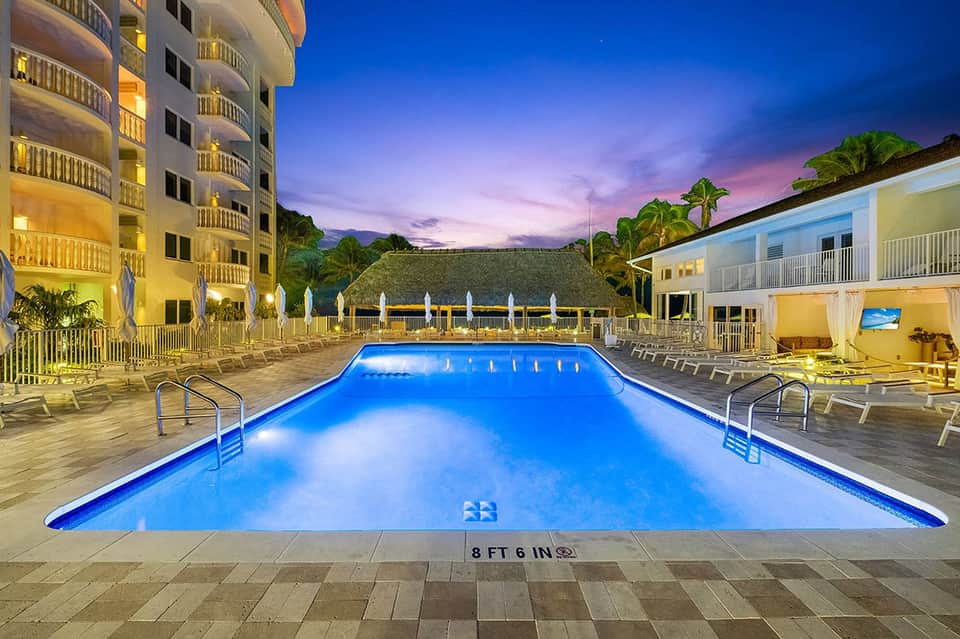 Resort pool at night with glowing blue water, hotel building, loungers, and lit umbrellas