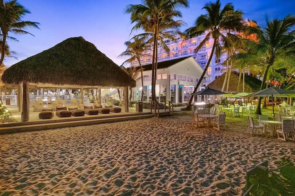 Beach lounge at dusk with tiki bar, palm trees, and hotel building lit up at night