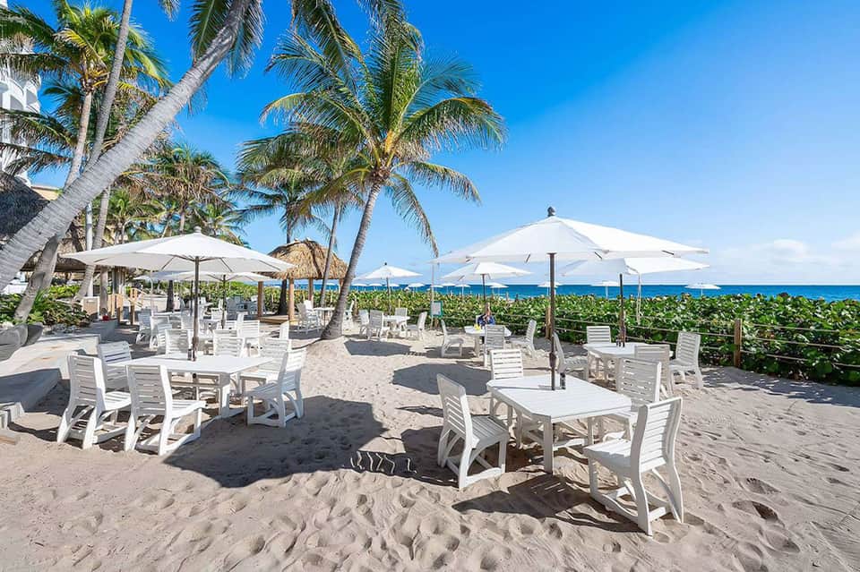 Beachfront dining area with white umbrellas, lounge chairs, palm trees, and ocean view