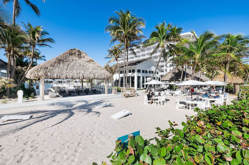 Beachfront resort with thatched-roof cabana, modern white building, palm trees, and beach seating