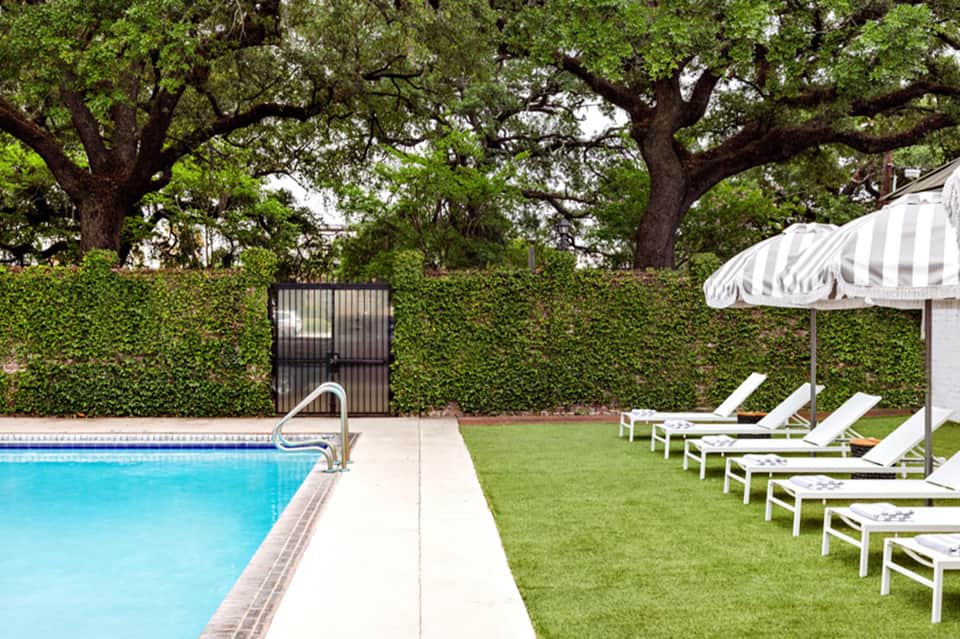Residential pool surrounded by manicured lawn, mature oak trees, white lounge chairs, and striped umbrella