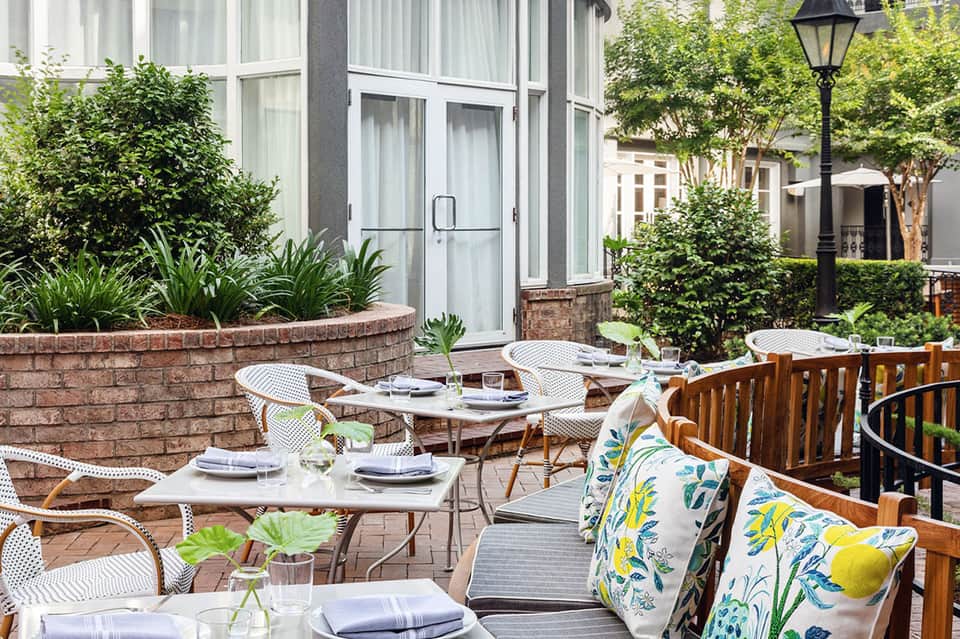 Outdoor courtyard dining space with white chairs, green landscaping, and brick planter walls