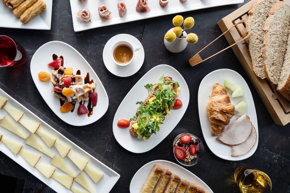 Fine dining breakfast spread with pastries, meats, fresh fruit, and espresso on dark table