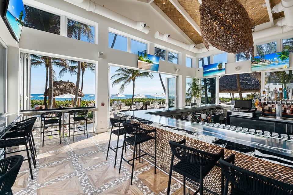 Beachfront bar with ocean views, palm trees, and tall windows opening to patio