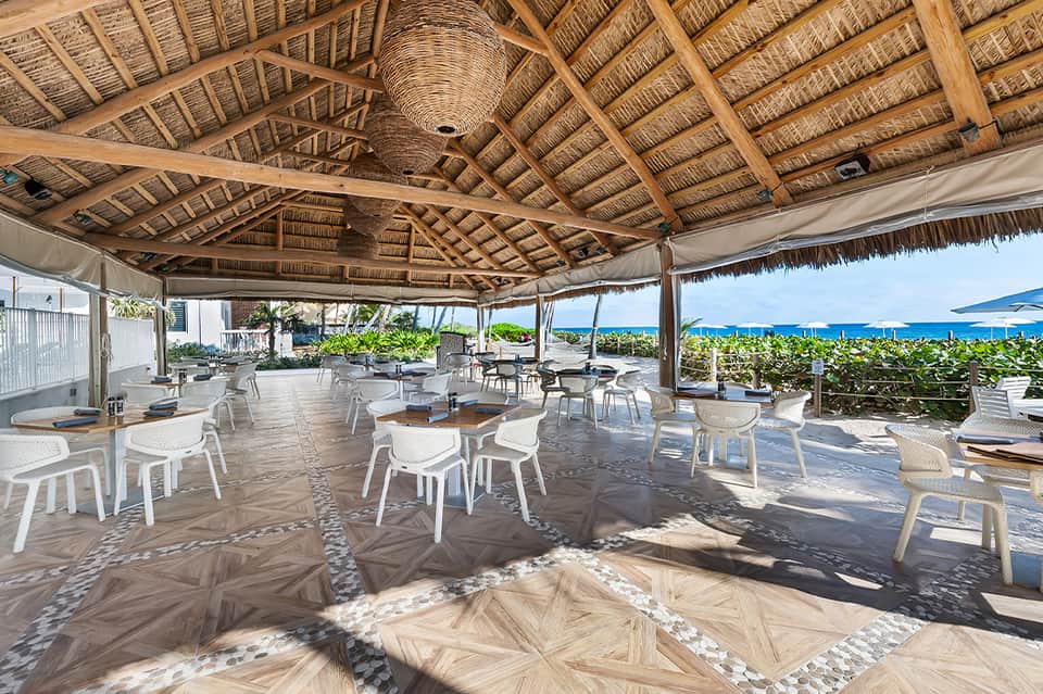 Upscale beachfront dining pavilion with thatch roof, white chairs, ocean view, and natural lighting