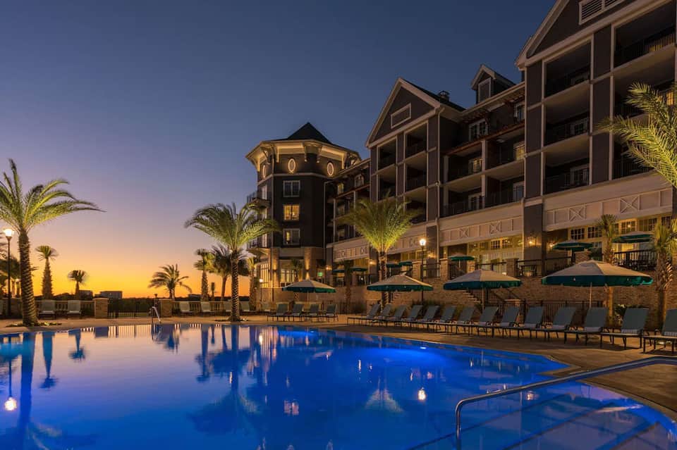 Illuminated beachfront resort at dusk with lit pool, palm trees, and waterfront buildings