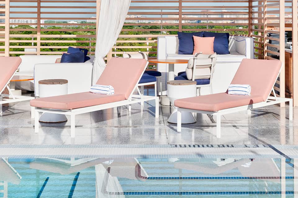 Poolside lounge area with pink cushioned loungers, white furniture, striped shade screen, and plush seating pods