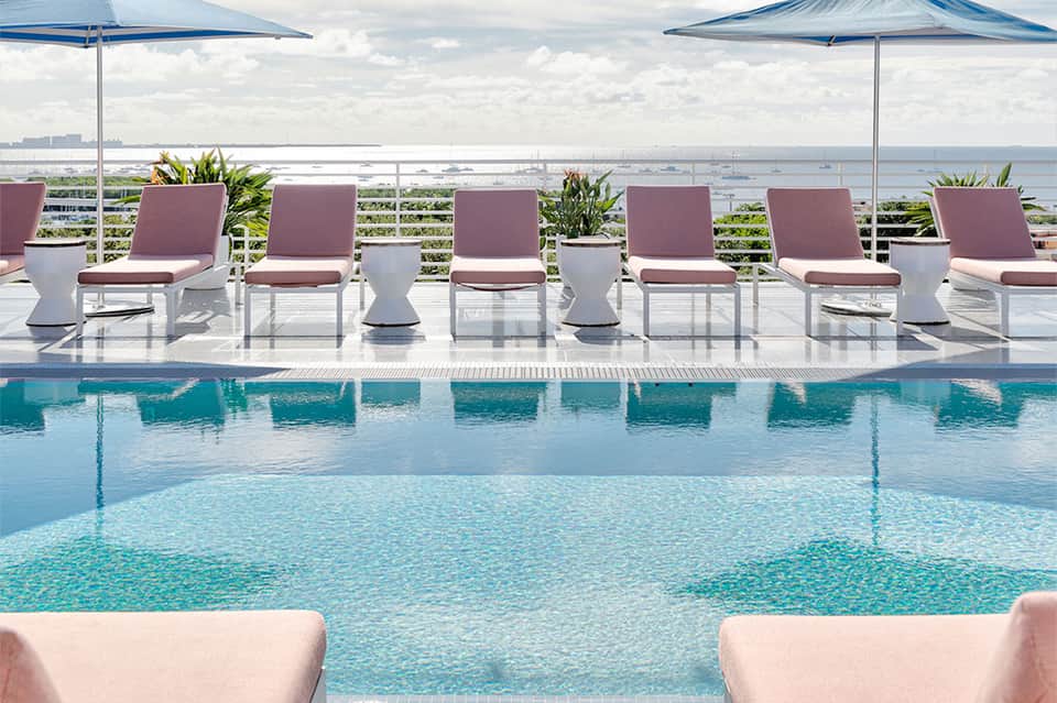 Resort rooftop pool overlooking ocean with white lounge chairs, blue umbrellas, and coastal views