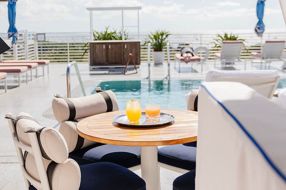 Oceanfront rooftop lounge with round wooden table, cream chairs, pool, and sea view beyond railing