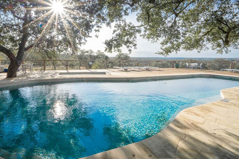 Resort pool with clear blue water, stone patio, and expansive landscape view under large shade tree