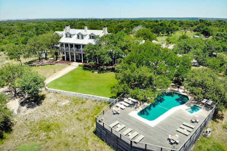 Aerial view of white manor house surrounded by green lawn and trees with pool deck below