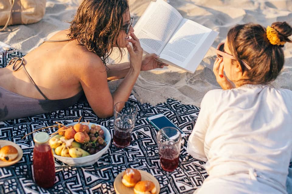 Two women reading and enjoying fruit and beverages on sandy beach with ocean view