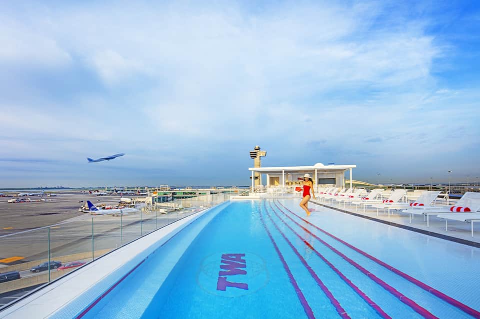 Rooftop swimming pool with lane markers overlooking airport runway, airplanes, and blue sky.