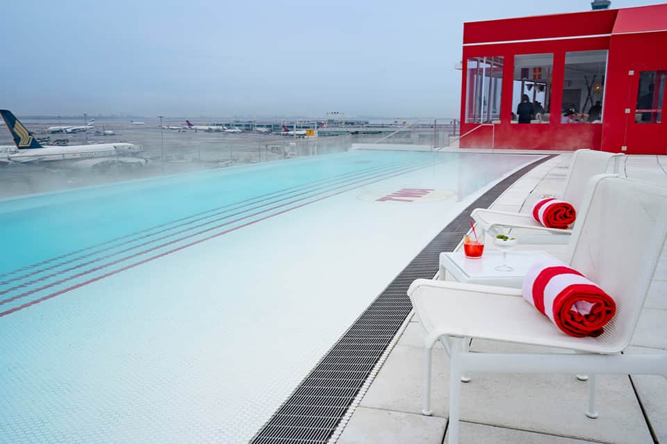 Rooftop lap pool with red cabana building, city airport views, and loungers with red towels