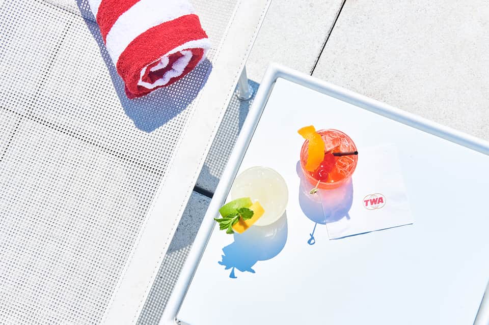 Colorful cocktails on white tray with TWA logo beside pool lounge chairs and red towel