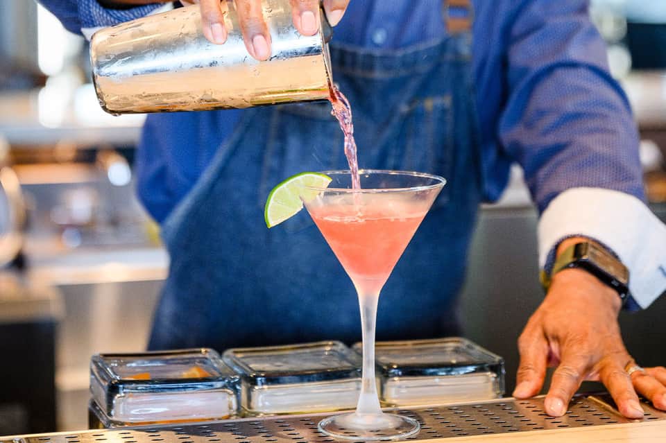 Bartender pouring coral-colored cocktail into martini glass with lime garnish