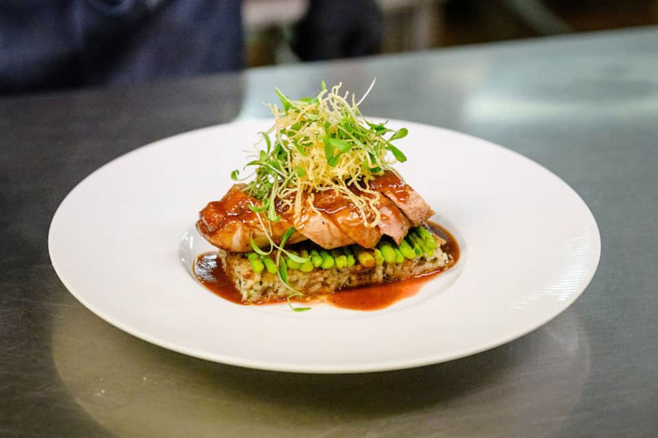 Gourmet plated duck dish with glazed protein, pea garnish, crispy noodles, and sauce on white plate