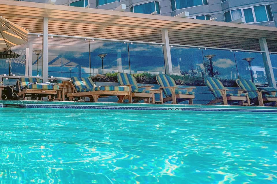 Resort pool with striped loungers, covered pergola, and ocean city view behind glass barrier