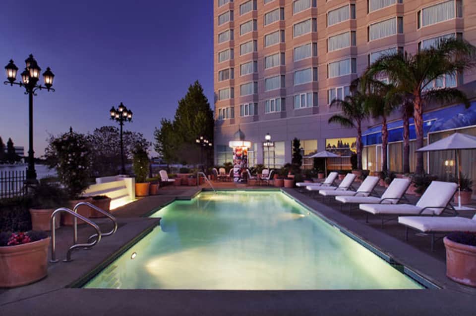 Illuminated rooftop pool at dusk with lounge chairs, palm trees, and brick building facade