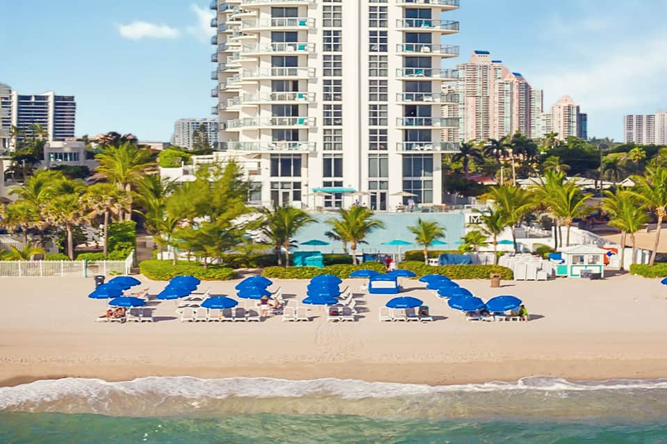 Beach scene with rows of blue umbrellas and lounge chairs fronting high-rise coastal resort buildings
