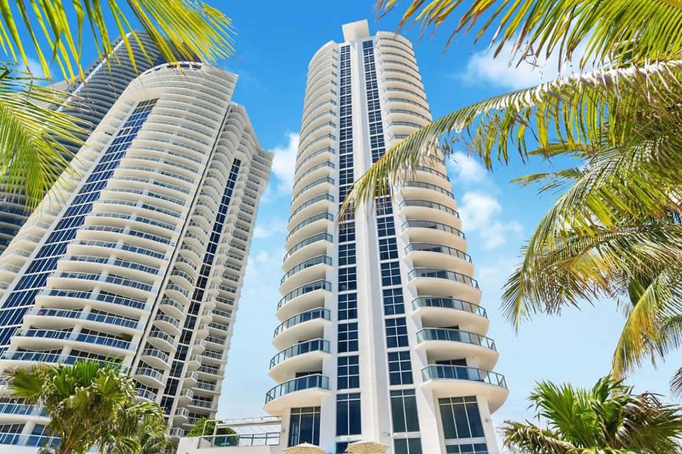 Modern high-rise residential towers with curved balconies framed by palm tree fronds against blue sky
