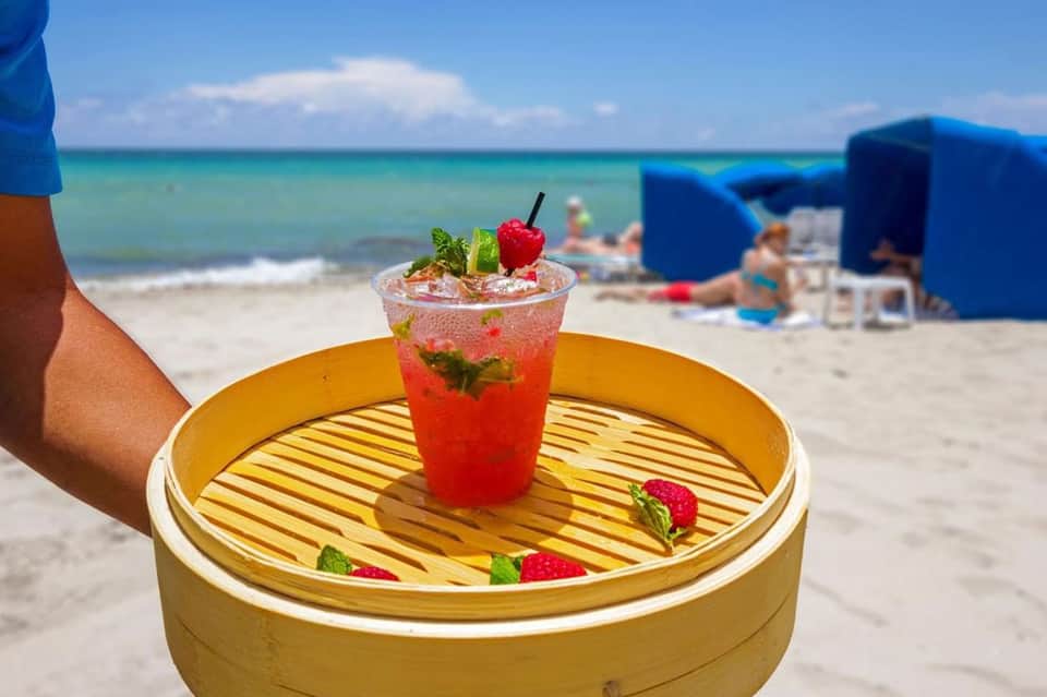 Red tropical cocktail on a wooden tray held at a beach with ocean in background