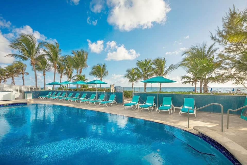 Curved resort pool with turquoise loungers, umbrellas, palm trees, and ocean view at horizon
