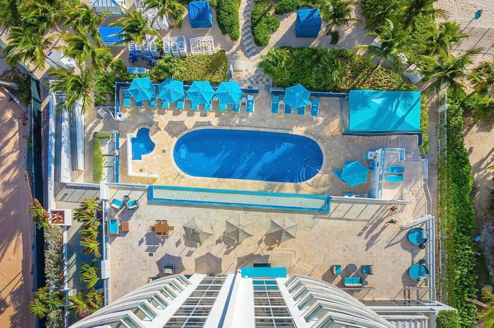 Overhead view of curved pool with blue umbrellas, lounge chairs, palm trees, and resort grounds
