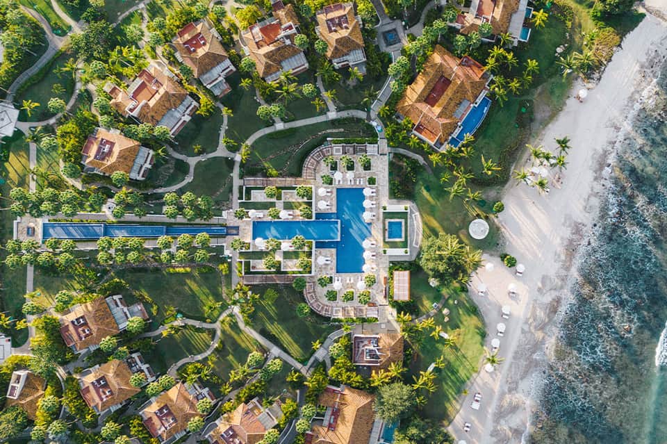 Aerial view of beachfront resort with curved blue pool, villas, gardens, and ocean