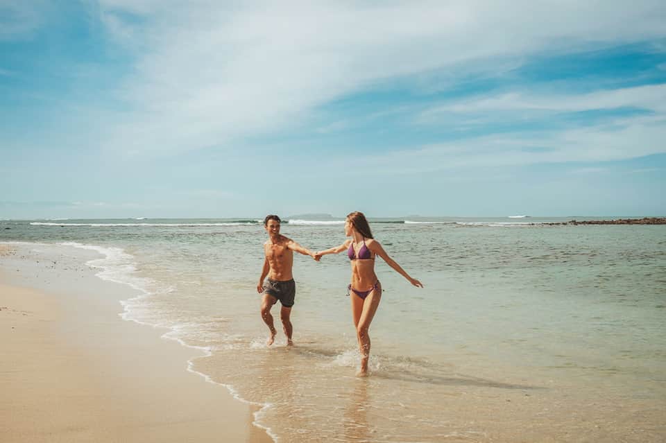 Couple running into ocean waves on sandy beach with clear blue water on sunny day