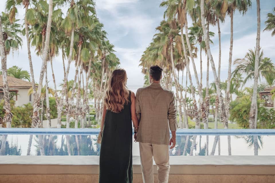 Couple holding hands overlooking a pool lined with palm trees and tropical landscaping