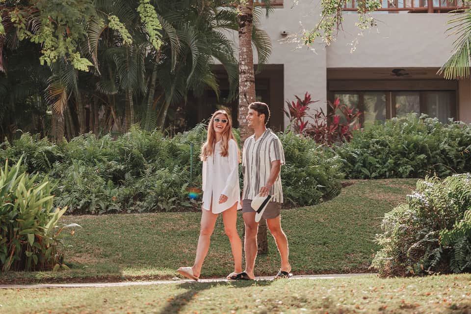 Couple walking through lush courtyard garden with tropical plants, palm trees, and white building architecture