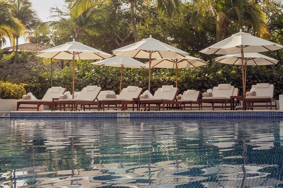 Resort pool with cream umbrellas, wooden loungers, palm trees, and calm water reflection
