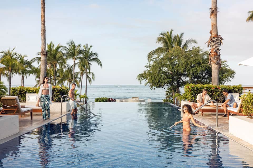 Infinity pool overlooking ocean with palm trees, guests, and coastal views