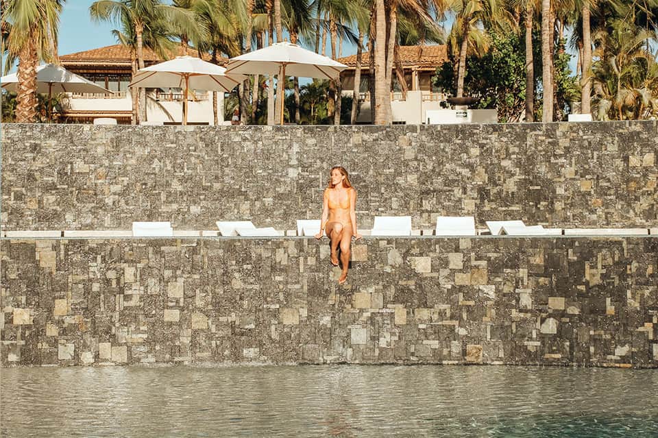 Woman in swimwear standing on stone waterfront wall with palm trees and buildings behind